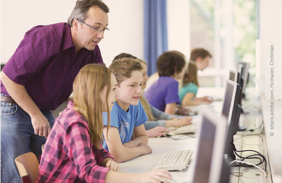 Bibox Sekundarstufe Ein Lehrer sieht drei Schülerinnen über die Schulter. Sie sitzen vor einem PC Bildschirm und arbeiten digital