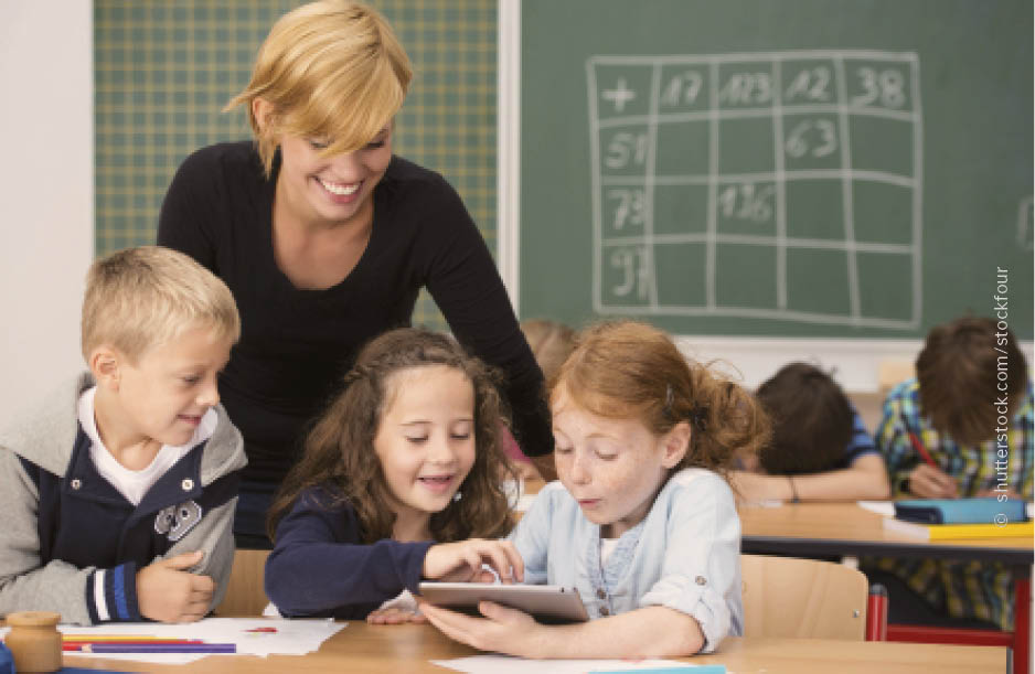 Bibox Volksschule Eine Lehrerin steht lächelnd im Klassenraum hinter drei Schülerinnen, die ein Tablet bedienen
